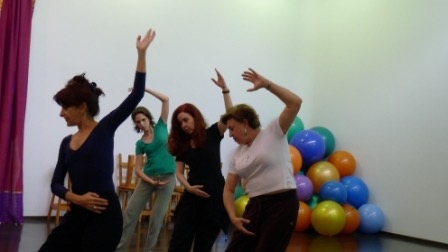 Um grupo de mulheres está participando de uma atividade de dança ou movimento em uma sala com bolas de exercício coloridas empilhadas ao fundo.