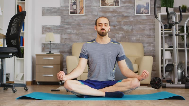 Um homem sentado em um tapete de ioga, praticando meditação mindfulness em uma sala de estar, com os olhos fechados e em uma postura relaxada.