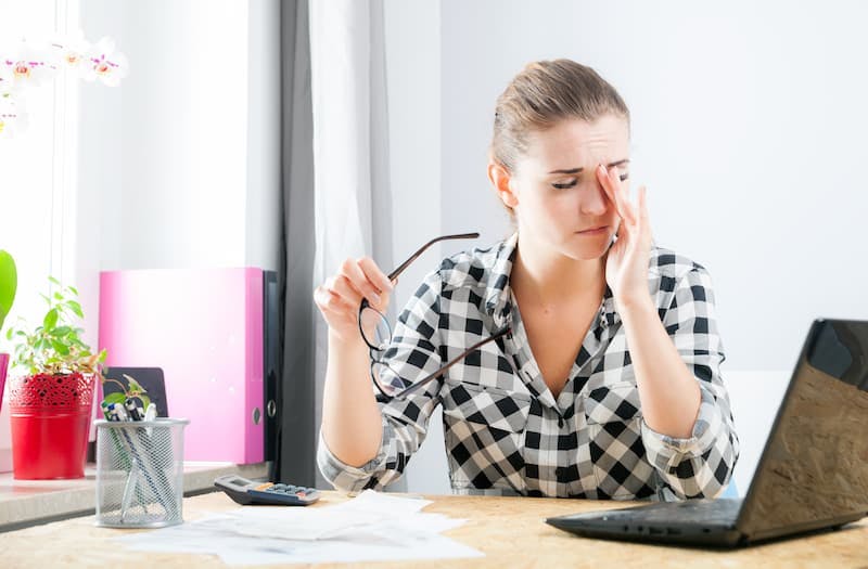 Uma mulher sentada à mesa de trabalho, aparentando cansaço, segura os óculos com uma mão enquanto esfrega os olhos com a outra. Em sua mesa, há um laptop, papéis, uma calculadora e alguns itens de escritório.