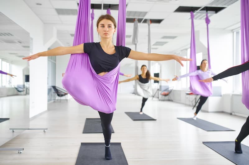 Sala de treinos de Air Yoga, com uma moça em foco fazendo uma posição de equilíbrio utilizando um pano lilás, roupa e tapete de yoga pretos