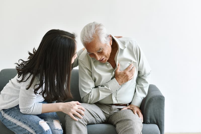 A imagem mostra um homem idoso sentado em um sofá, claramente em situação de desconforto ou dor. Ele está segurando o peito com a mão direita, sugerindo que pode estar sofrendo um ataque cardíaco ou algum outro problema relacionado ao coração. Ao lado dele, uma jovem mulher está inclinada em sua direção, demonstrando preocupação e oferecendo apoio. Ela parece estar tentando ajudar ou conversar com ele para entender melhor a situação. O ambiente é simples e claro, com um fundo branco que destaca os dois indivíduos no sofá.