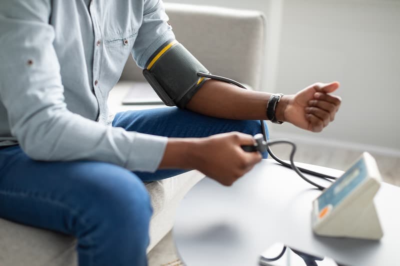 A imagem mostra uma pessoa medindo a pressão arterial em casa. Ela está sentada em um sofá, usando uma camisa de manga comprida azul clara e calça jeans. O braço esquerdo está apoiado e possui uma braçadeira de pressão arterial inflada. A pessoa está segurando a bomba de borracha usada para inflar a braçadeira com a mão direita. Em uma mesa à frente, há um monitor digital de pressão arterial que está conectado à braçadeira.